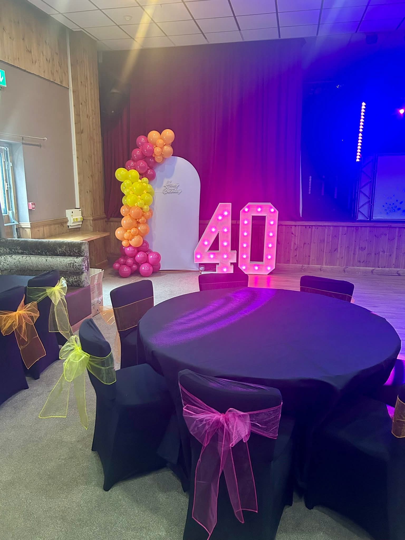 Black table covers with pink, orange and yellow chair sashes, an orange and pink balloon column and light-up 40 numbers